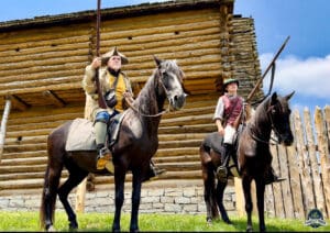 Two men on horses in front of a log cabin.